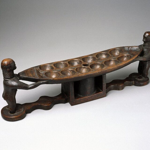 The object is a boat-shaped board with 12 playing cups and one triangular "capture" cup at either end. The playing area stands on a rectangular box which originally held playing seeds. The two ends are supported by a pair of male figures with ringed necks. The proper left leg of one of the figures is missing. English: Mancala is one of the world's oldest games and is widely played in Africa. This board, like most, has twelve holes and two large cups to hold each player's game pieces. The figurative carvings, however, are rare, and the board's boat shape reflects the importance of canoes to the Bidjogo. Mancala reflects ideas about how society is organized: the board may represent either the village or the universe; the holes are called "houses" or "villages"; and the playing pieces, which are moved around the board, are called "men," "wives," "children," or "cattle." - Online Collection of Brooklyn Museum; Photo: Brooklyn Museum, 22.239_SL1.jpg